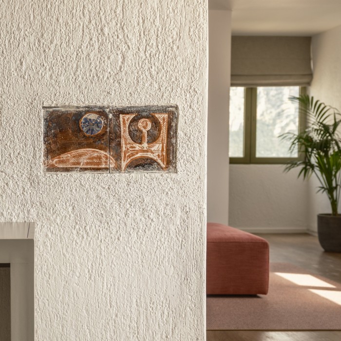 Photo of a roughly plastered pale wall in a hotel room, with a rectangular, textured tile embedded into the wall, featuring an bold, abstract piece of art